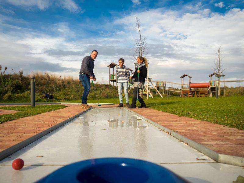 Drei Personen spielen Minigolf auf einem Außenplatz bei bewölktem Himmel.