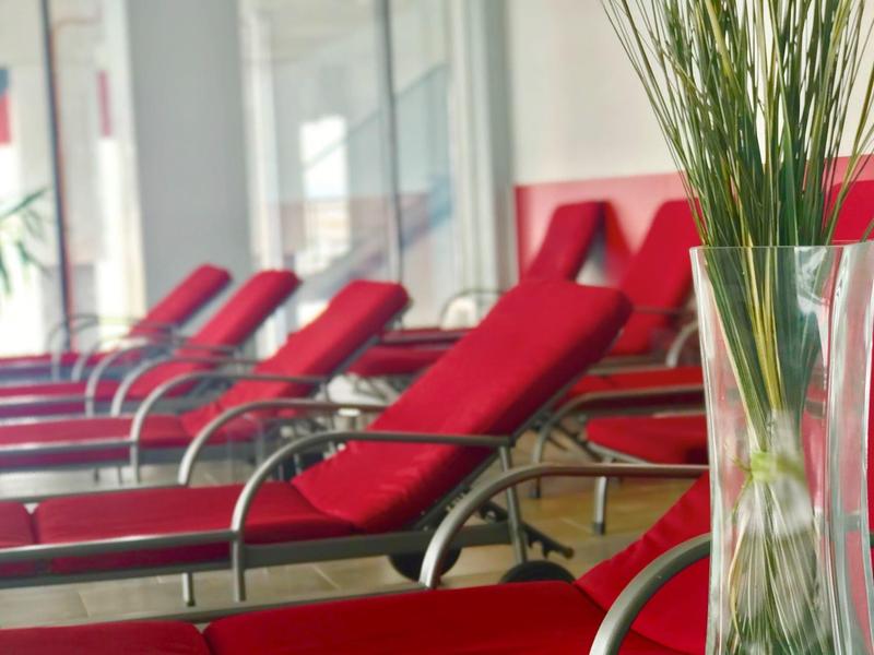 Rote Liegestühle in einem hellen Innenbereich, im Vordergrund eine Vase mit grünen Gräsern.