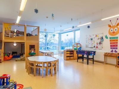 Heller, kinderfreundlicher Raum mit Spielbereichen, Tischen und bunter Dekoration für Betreuung.