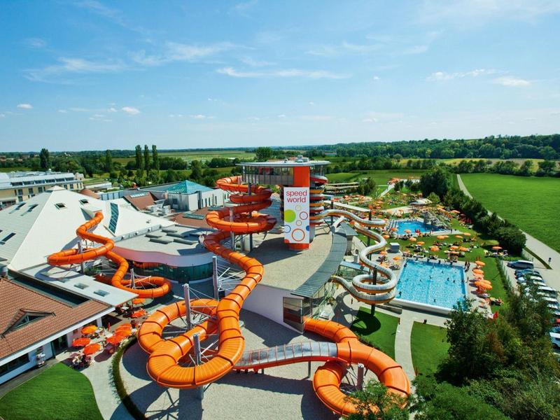 Wasserpark mit großen orangefarbenen Wasserrutschen und Schwimmbecken neben grüner Landschaft.