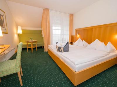 Hell eingerichtetes Hotelzimmer mit Doppelbett, Schreibtisch und Stuhl vor Fenster mit Vorhängen.