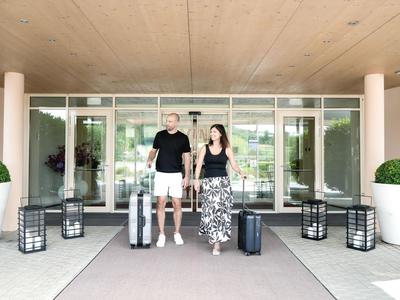 Ein Paar betritt mit Koffern den Eingangsbereich eines Hotels mit Glasfassade und Pflanzen.
