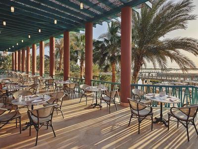 Terrasse couverte avec tables et chaises à côté de palmiers et vue sur la mer au coucher du soleil.