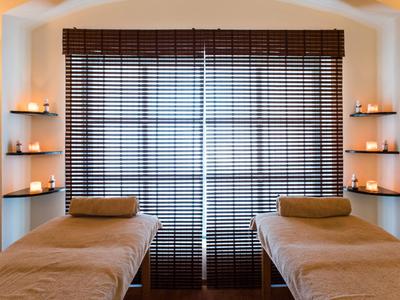 Zwei Massageliegen mit braunen Bezügen vor großen Fensterläden und vier eingerahmten Kerzenhaltern.