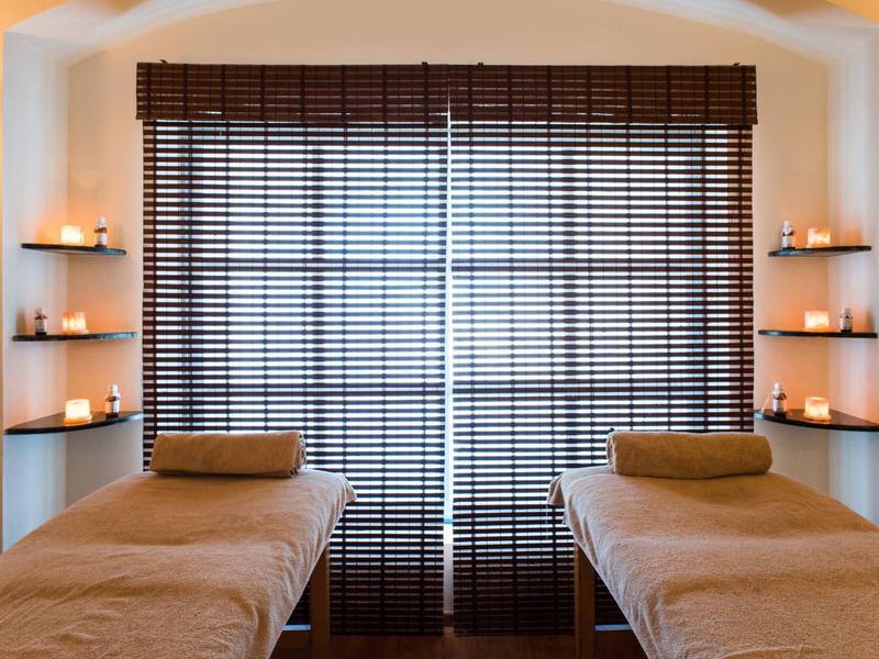 Zwei Massageliegen mit braunen Bezügen vor großen Fensterläden und vier eingerahmten Kerzenhaltern.