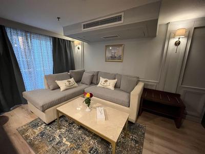 Helle Lounge mit grauem Ecksofa, hellem Couchtisch und großem Fenster mit Vorhängen.