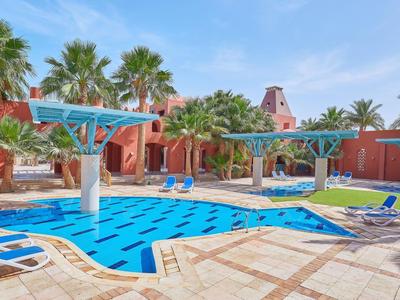 Freizeitzentrum mit blauem Pool, weißen Liegestühlen, Palmen und roten Gebäuden bei Sonnenschein.