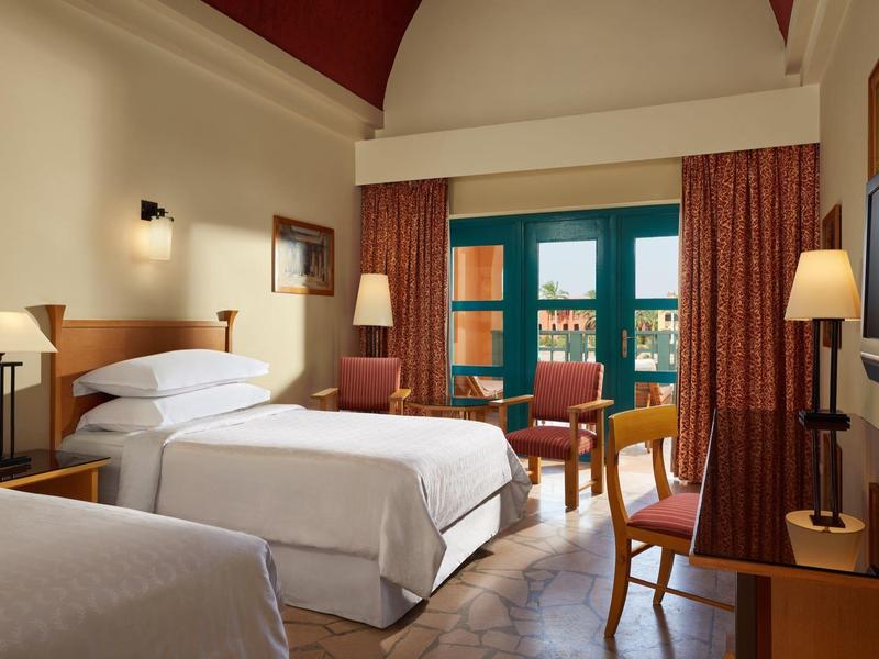 Zwei Einzelbetten in einem Hotelzimmer mit Holzmöbeln, Vorhängen und grünem Fensterrahmen.