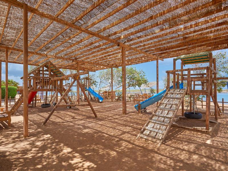 Überdachter Spielplatz mit Klettergerüsten, Schaukeln und Rutschen aus Holz bei Sonnenschein.