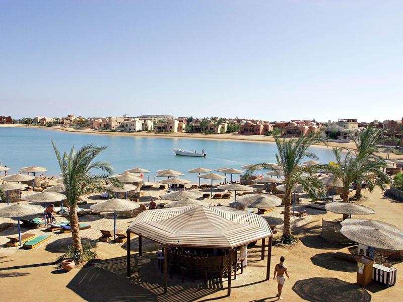 Strand mit Sand, Sonnenschirmen, Palmen und blauem Wasser unter klarem Himmel.