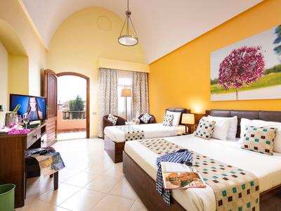 Heller Hotelraum mit drei Einzelbetten, gelber Wand, Balkon und Fernseher auf dunklem Möbelstück.