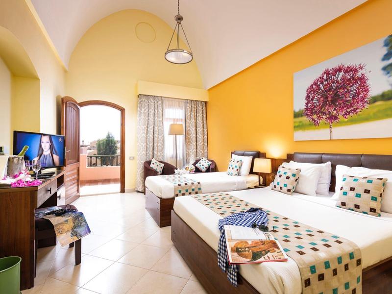 Heller Hotelraum mit drei Einzelbetten, gelber Wand, Balkon und Fernseher auf dunklem Möbelstück.