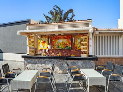 Zona exterior vacía de cafetería con mesas, sillas y una barra colorida al fondo.