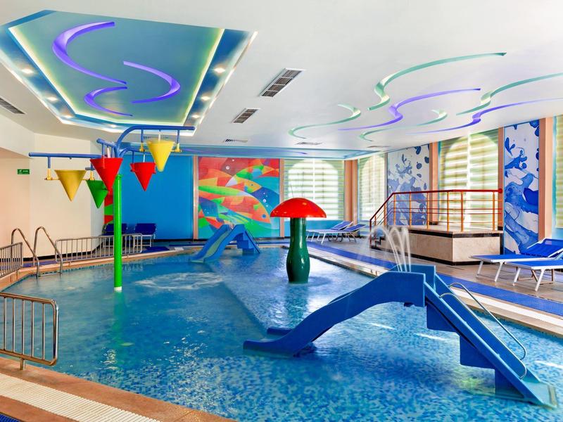 Piscine intérieure colorée avec toboggan et chaises longues près de grandes fenêtres.