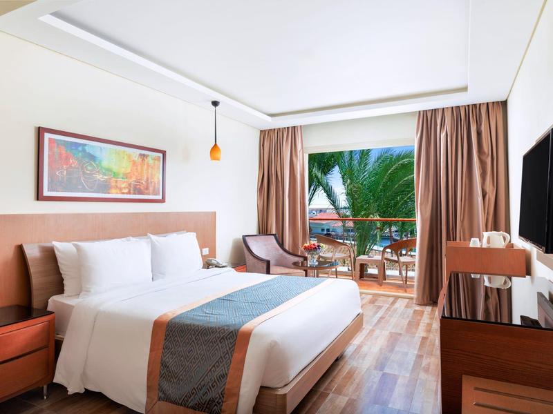 Bright hotel room with large bed, wooden furniture, and balcony overlooking palm trees.