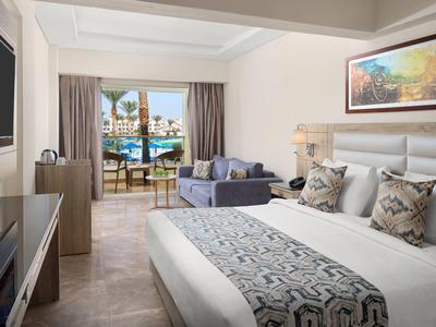 Modern hotelkamer met groot bed, bank en uitzicht op zwembad en terras.