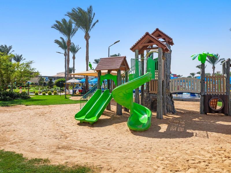 Speeltuin met groene glijbaan en palmbomen in zonnig buitengebied van een resort.