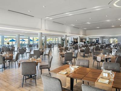 Ampia sala da pranzo moderna con tavoli in legno, sedie grigie, ambiente luminoso e grandi finestre.