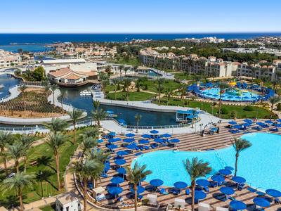 Grande piscina dell'hotel con lettini blu e palme, circondata da edifici dell'hotel e vista sul mare.