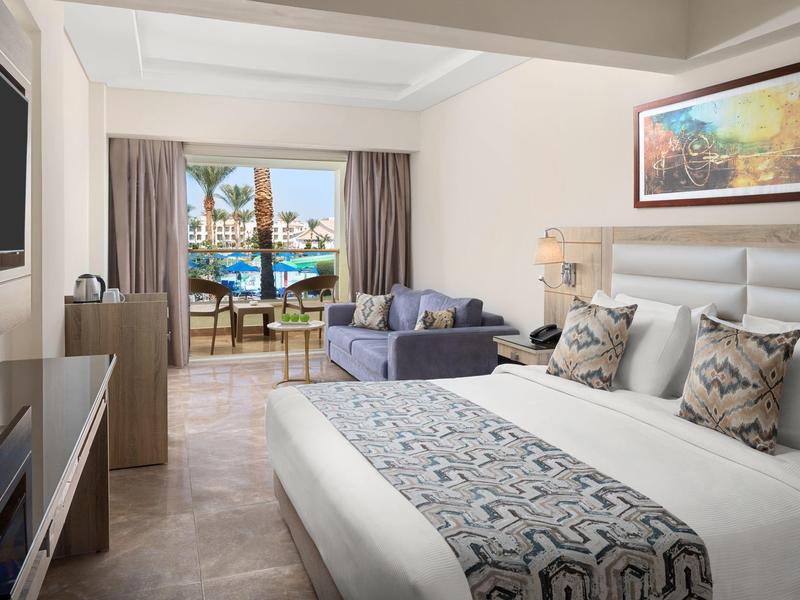 Chambre d'hôtel élégante avec lit double, canapé, TV, balcon donnant sur piscine.