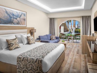 Modern hotel room with bed, sofa, and terrace overlooking garden and pool