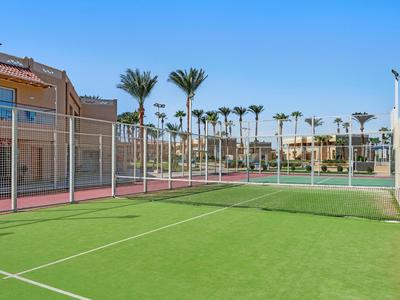 Empty tennis court with green and red surface, surrounded by buildings and palm trees under a blue sky.