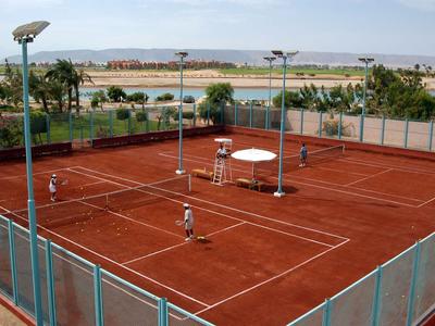 Varios campos de tenis de arcilla roja rodeados de cercas verdes en una ubicación soleada.
