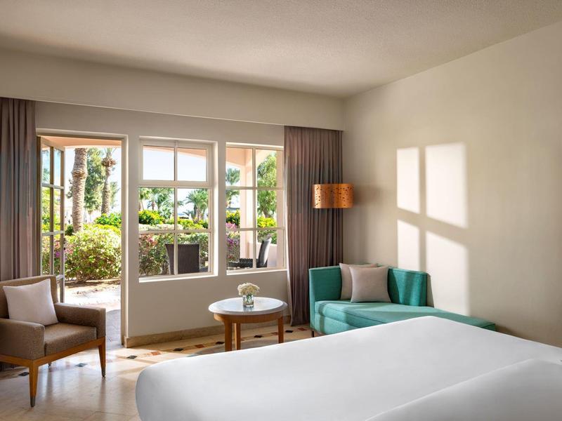 Helles Schlafzimmer mit Doppelbett, Sofa, Stühlen und Glasfenstern zum grünen Balkon.