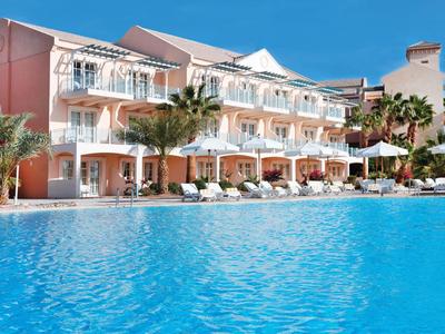 Grande hotel con piscina blu e lettini prendisole sotto un cielo azzurro.