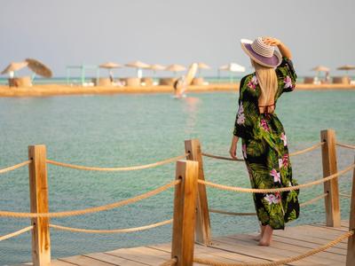 Femme portant un chapeau et une robe fleurie debout sur un quai avec vue sur l'eau et des transats en arrière-plan.