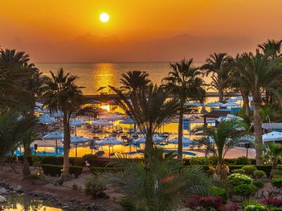 Atardecer sobre un resort de playa con palmeras y tumbonas junto al agua.