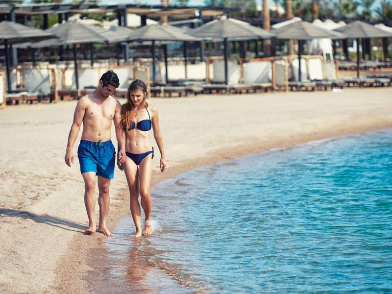 Ein Paar geht am Strand entlang, mit Sonnenschirmen und Liegestühlen im Hintergrund.