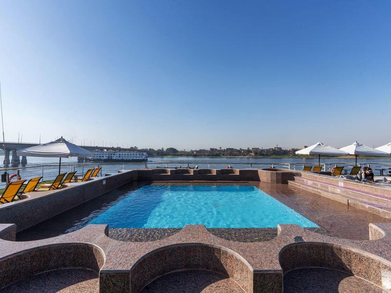 Piscina all'aperto con lettini vicino a un lago in una giornata di sole.
