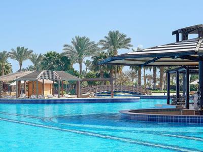 Una grande piscina con acqua azzurra limpida davanti a palme e un bar coperto nell'area dell'hotel.