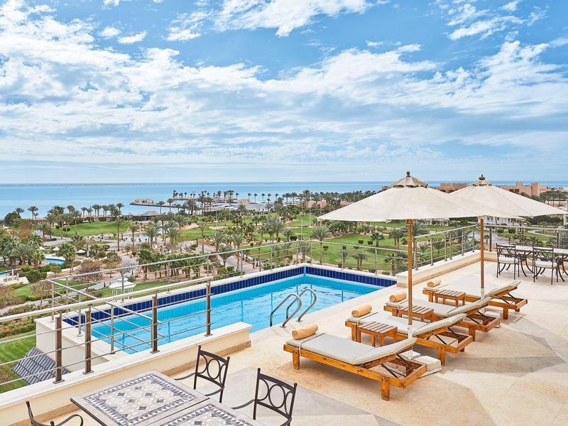 Terraza con piscina, tumbonas y sombrillas con vista al mar y palmeras bajo un cielo nublado.