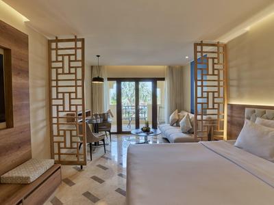 Modern hotel room with large bed, seating area, and balcony doors overlooking plants.