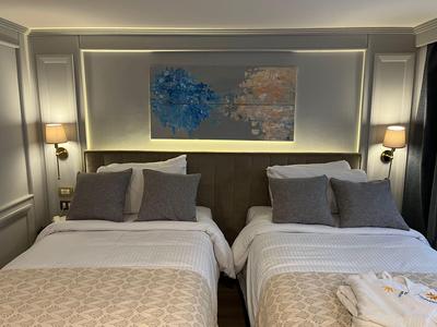 Camera d'hotel con due letti singoli, cuscini grigi, coperte beige e opera d'arte murale sopra la testiera.