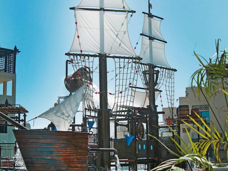 Een houten piratenschip in een hotelzwembad met palmbomen en blauwe hemel.