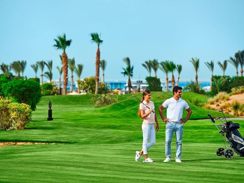 Pár hrající golf na zeleném hřišti s palmami a modrou oblohou v pozadí.