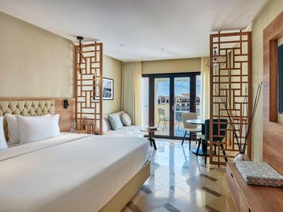 Chambre d'hôtel lumineuse avec grand lit, deux fauteuils et terrasse avec vue sur la mer.