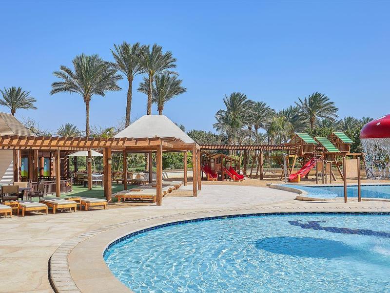 Piscine de l'hôtel avec jeu d'eau et espaces assis sous des pavillons en bois par temps ensoleillé.