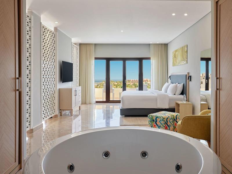 Habitación de hotel luminosa con cama cómoda, TV, terraza y bañera independiente en primer plano.