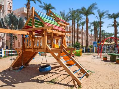Parque infantil con tobogán de madera, columpios de neumáticos y escalera rodeado de palmeras en arena.