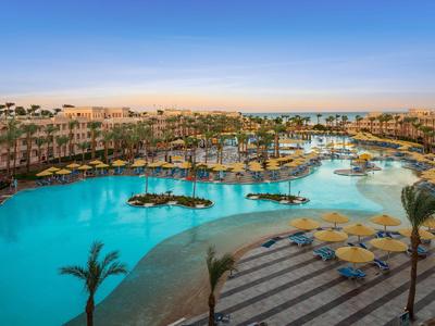 Large hotel complex with pool area, palm trees, and sun loungers under clear sky.