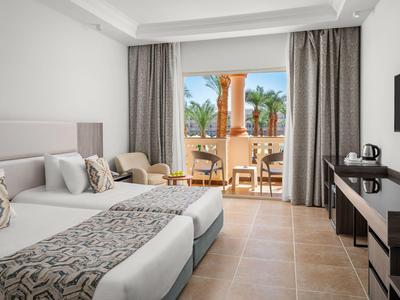 Habitación de hotel luminosa con dos camas y balcón con vistas a palmeras y columnas.