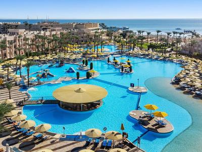 Großer Hotelpool mit Palmen, Sonnenschirmen und Meerblick bei blauem Himmel.