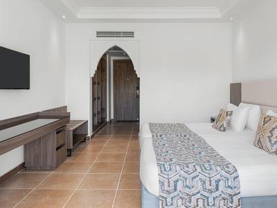 Habitación de hotel luminosa con cama doble, mesa, televisor y arco hacia el pasillo.