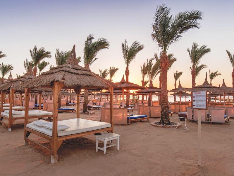 Playa con camas de sombra de palma y tumbonas al atardecer en un resort hotelero.