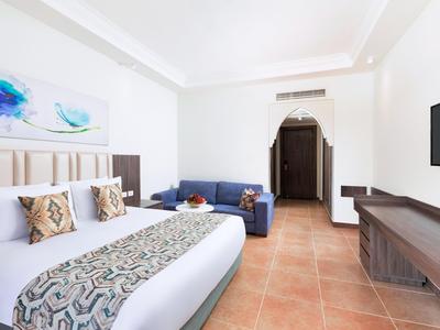 Lichte hotelkamer met groot bed, bank, tv en tegelvloer.