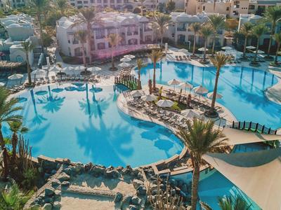 Grande piscina dell'hotel con palme, lettini e edifici circostanti sotto un cielo azzurro.